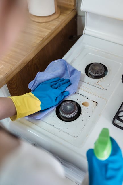 Close-up of a person wearing yellow and blue rubber gloves, using a blue cleaning cloth to scrub the stovetop of a white electric oven in a domestic kitchen. The stovetop features two black burners, o