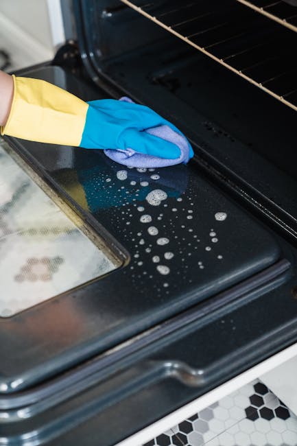A woman wearing a white t-shirt, yellow and blue cleaning gloves, is kneeling in a modern kitchen, cleaning the black ceramic oven interior with a spray bottle and a blue cloth. The oven door is open,